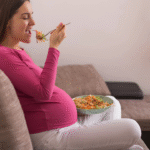 mulher grávida sentada no sofá da casa comendo uma alimentação saudável na gestação